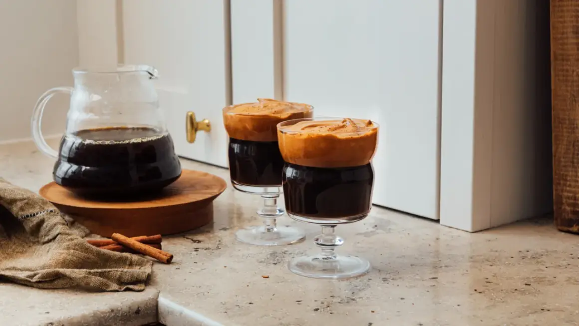 Två glas med mörkt, kallt kaffe toppat med fluffigt skum. En karaff med kaffe och kanelstänger står bredvid.