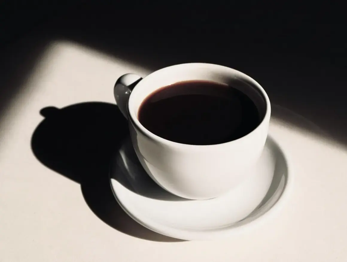 En vit kaffekopp med mörkt kaffe, belyst av solljus som skapar dramatiska skuggor. Hur påverkar kaffe blodtrycket?
