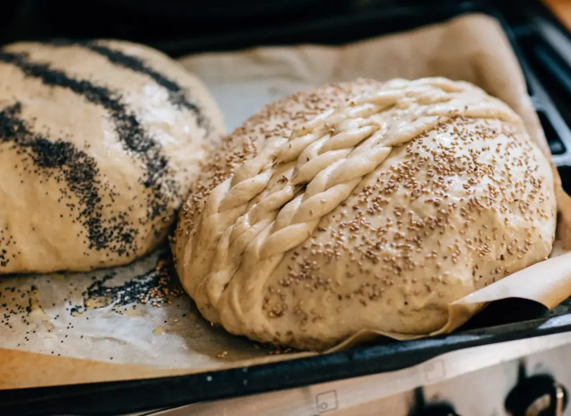 Två nybakade bröd på bakplåtspapper, ett toppat med vallmofrön och ett med sesamfrön och flätat mönster. Perfekta fika idéer!
