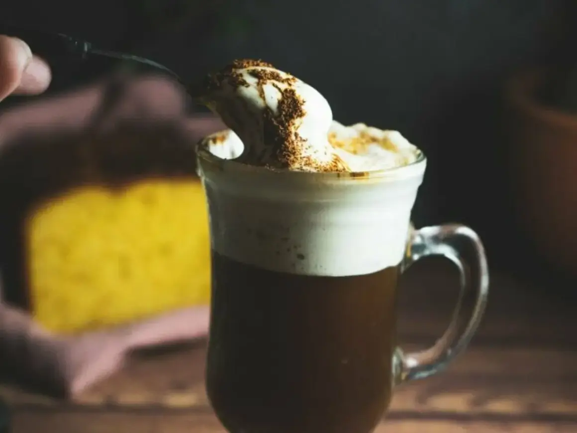 En sked med vispad grädde och kanel toppar en varm kaffe Karlsson utan Baileys. I bakgrunden syns en gul kaka.