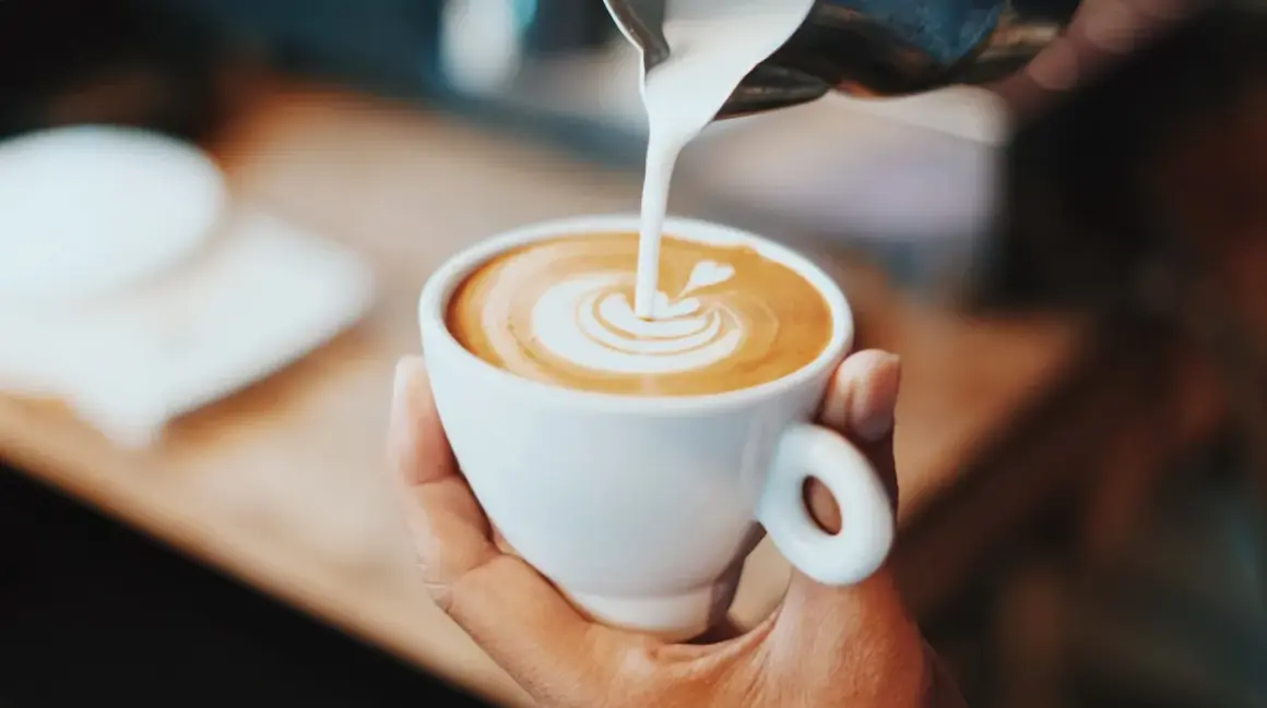 En hand håller en vit kopp med eritreansk kaffe, där mjölk hälls i för att skapa lattekonst.