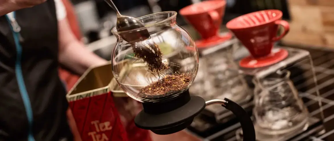 En sked lägger teblad i en glaskaraff, redo för att brygga te utan koffein. Röda kaffefilter står i bakgrunden.