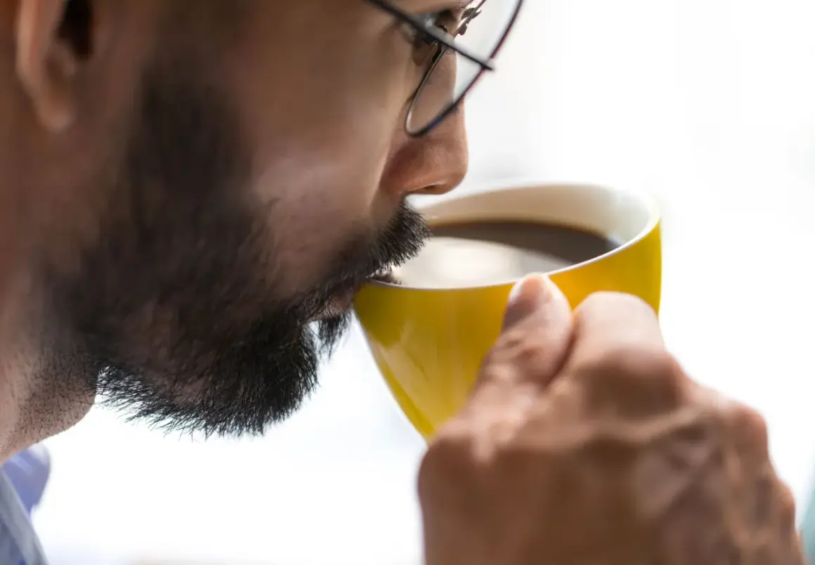 Man med glasögon dricker kaffe ur en gul mugg. Koffein ger energi, motverkar adenosin.