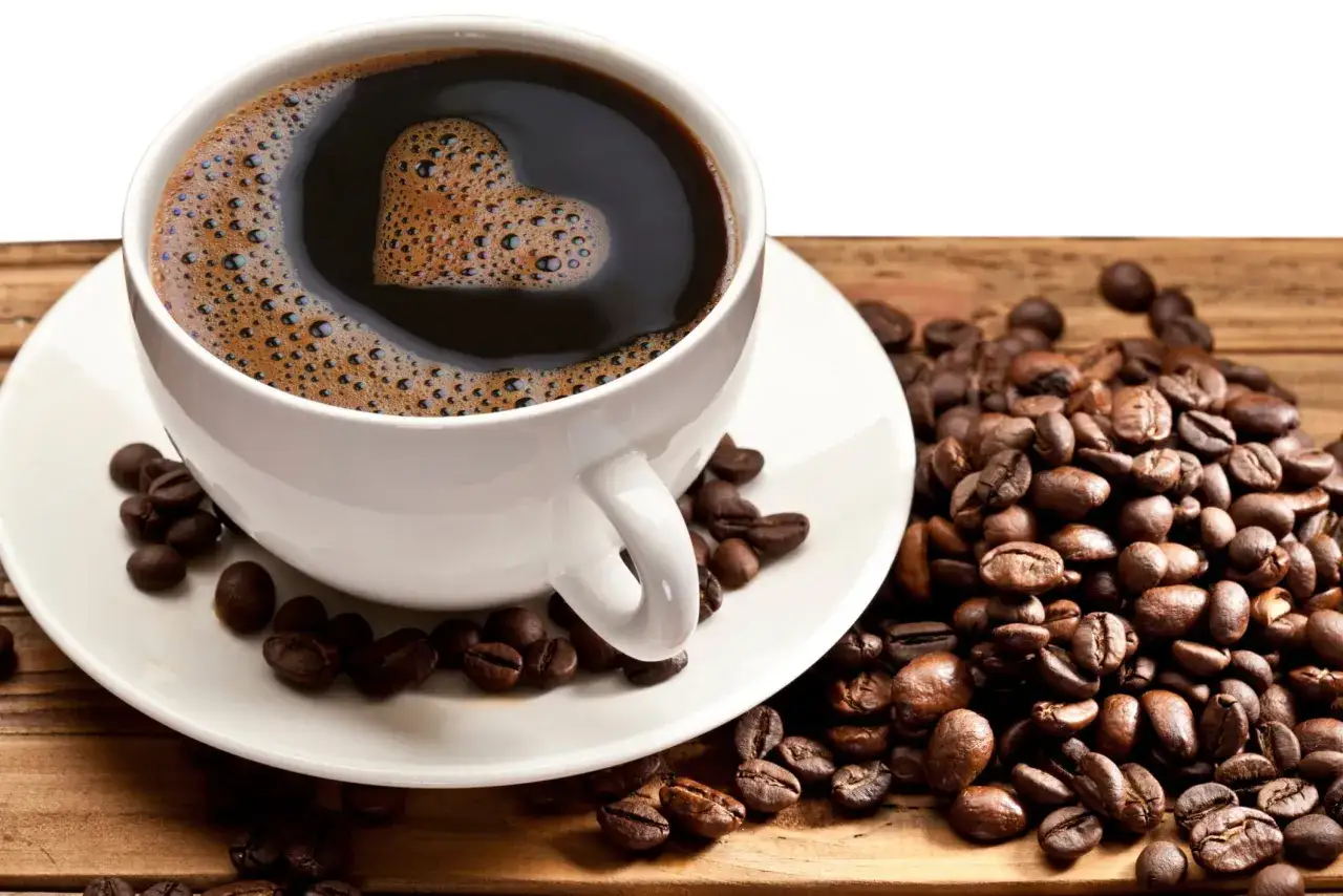 En vit kopp med kaffe där skummet bildar ett hjärta. Runt koppen ligger rostade kaffebönor, en påminnelse om att njuta av livet trots risker som cancer.