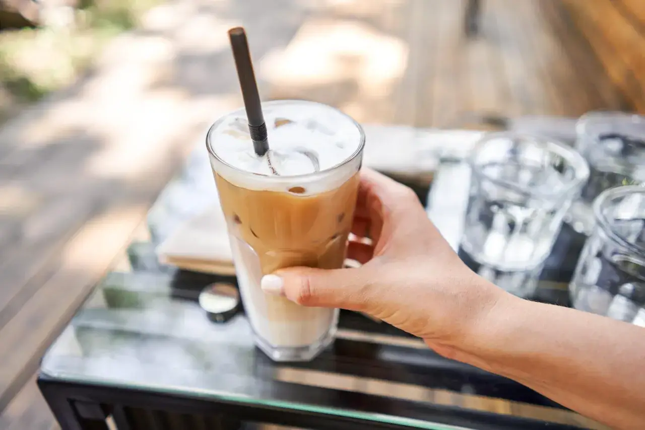 En hand håller ett glas iskaffe med skummig topp och en mörkbrun sugrör. Perfekt för ett iskaffe recept en varm sommardag.