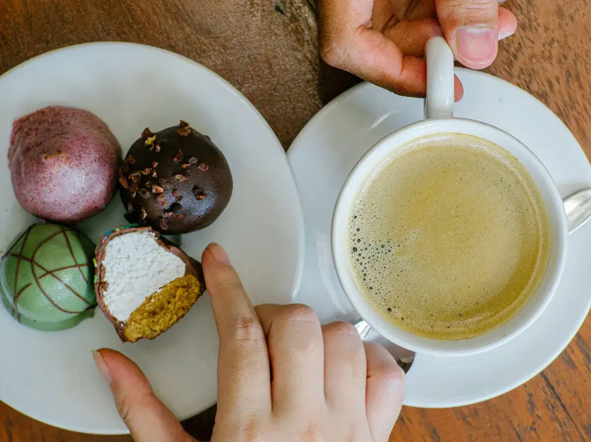 En kopp svensk kaffe med skummig yta bredvid en tallrik med färgglada bakelser. En hand sträcker sig efter en av bakelserna.