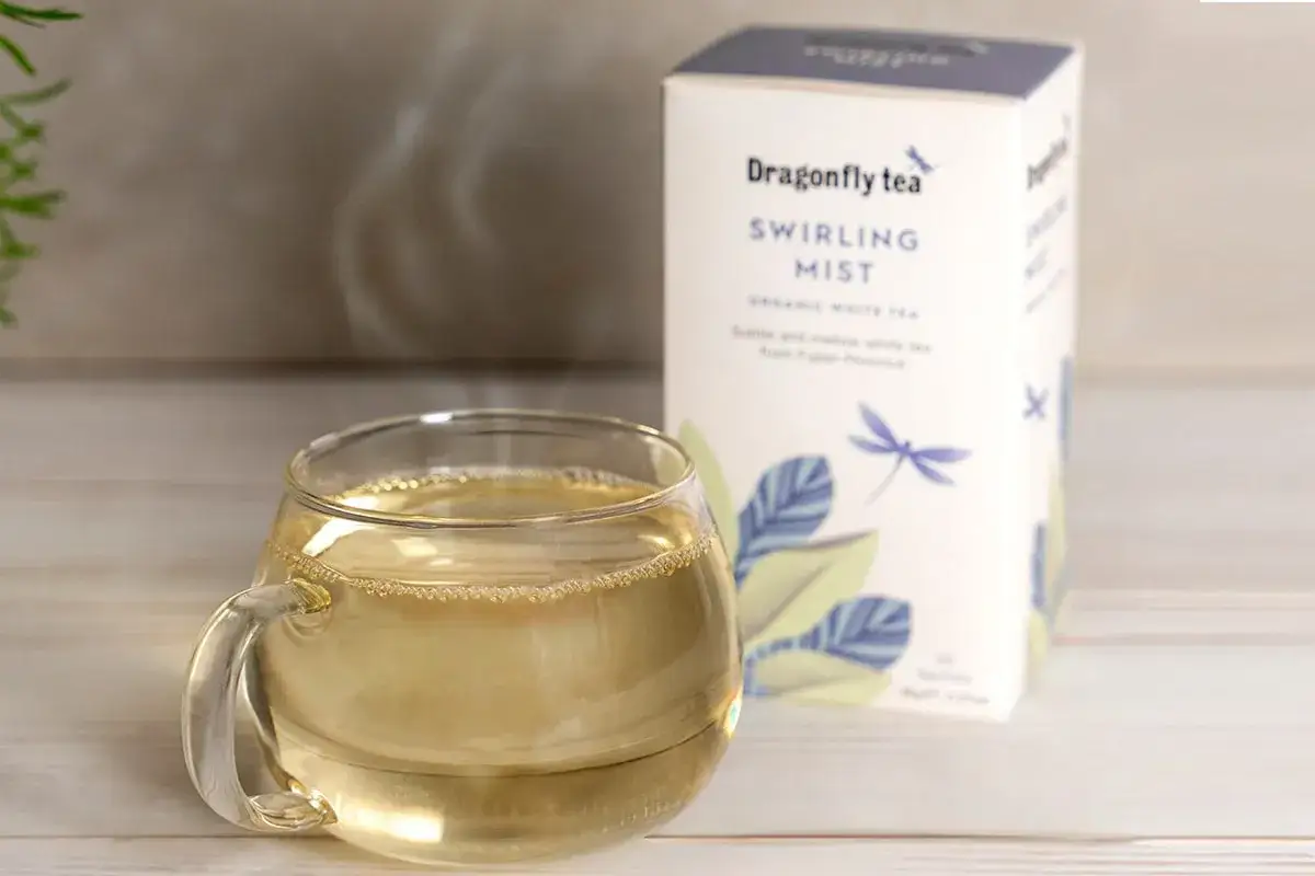 En kopp ångande vitt te bredvid en låda Dragonfly Tea Swirling Mist. Vitt te nyttigt för en lugn stund.