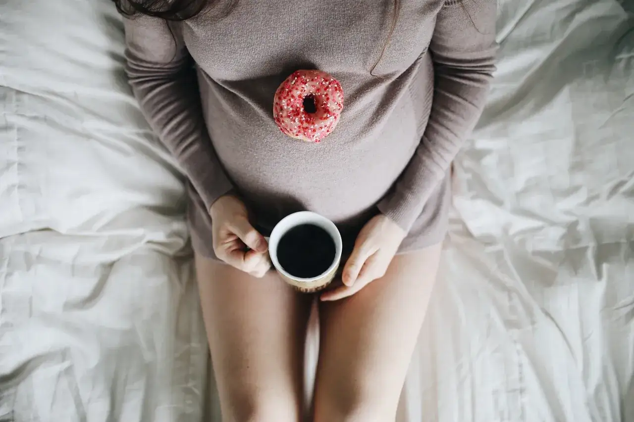 Gravid kvinna med en donut på magen och en kaffekopp i handen. Frågan 