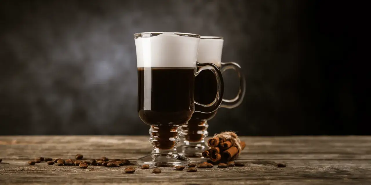 Två irish coffee utan farinsocker med krämigt skum, kaffebönor och kanelstänger på träbord.