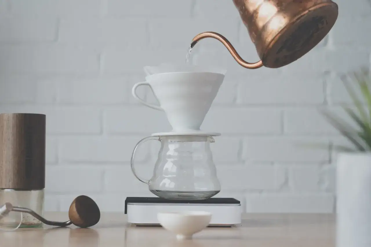 Vatten hälls från en kopparkanna över ett filter ner i en glaskaraff, för att göra dropp kaffe. En kaffekvarn står bredvid.