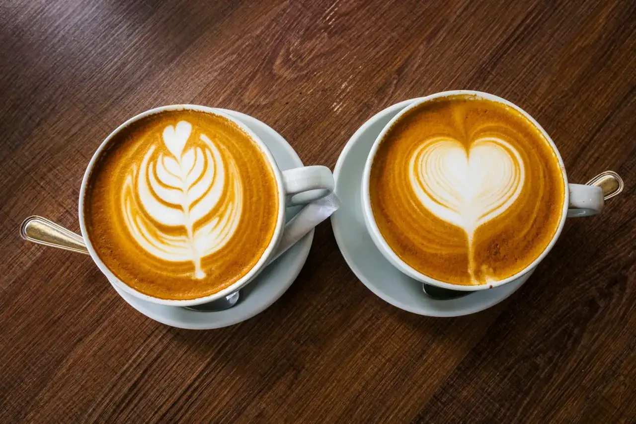 Två kaffekoppar med latte art på träbord. En med ett lövmönster, den andra med ett hjärta. En visuell skillnad mellan latte och cappuccino.