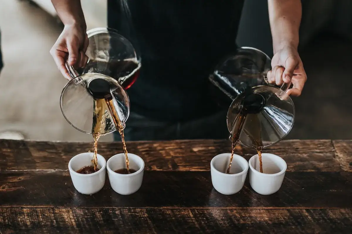 Två personer häller kaffe från glasbehållare till små vita koppar. En perfekt pour over-upplevelse.
