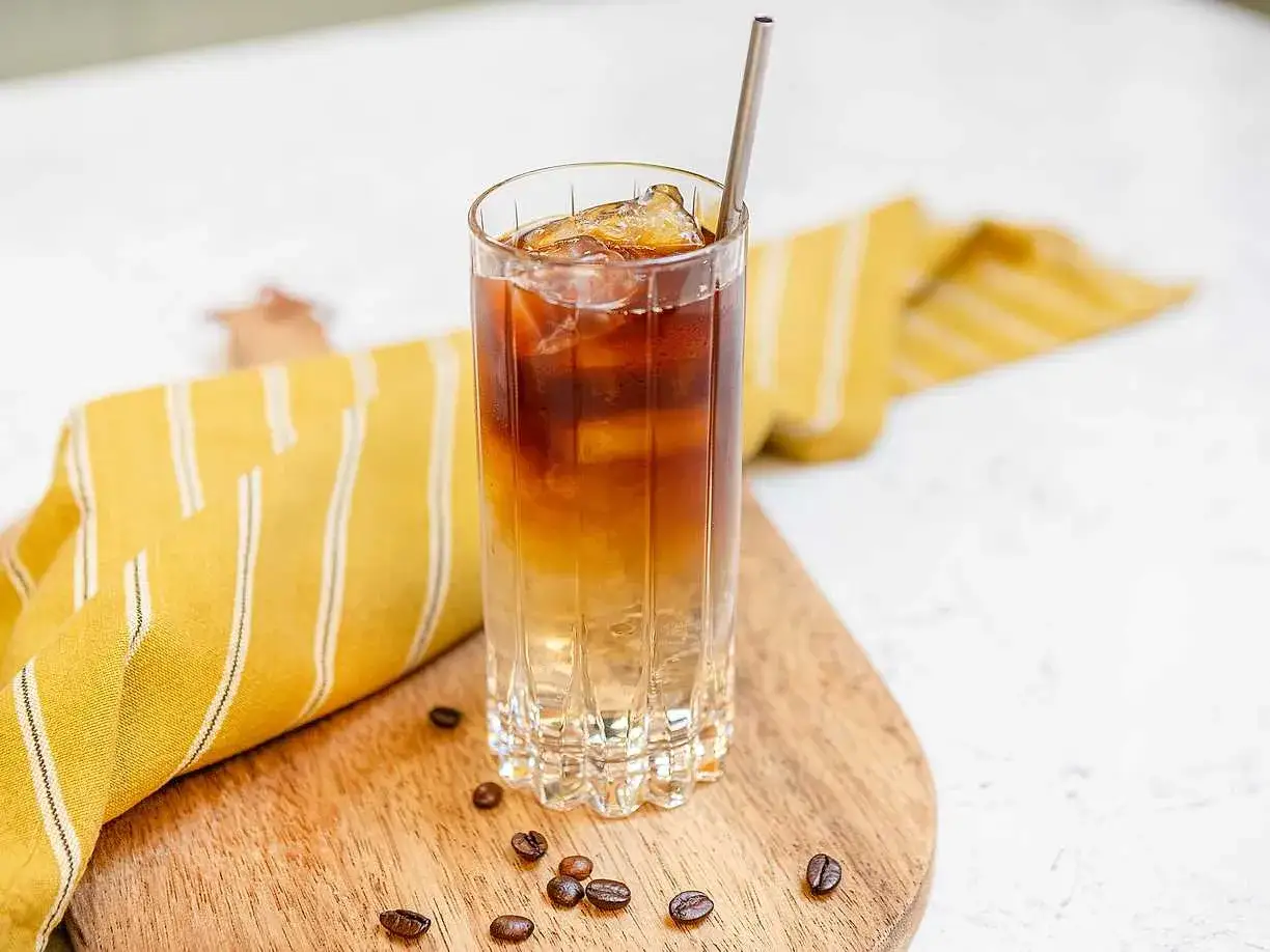 Ett glas med kaffe tonic, is och en metallhalm. En gulrandig servett och kaffebönor ligger på en träskärbräda.