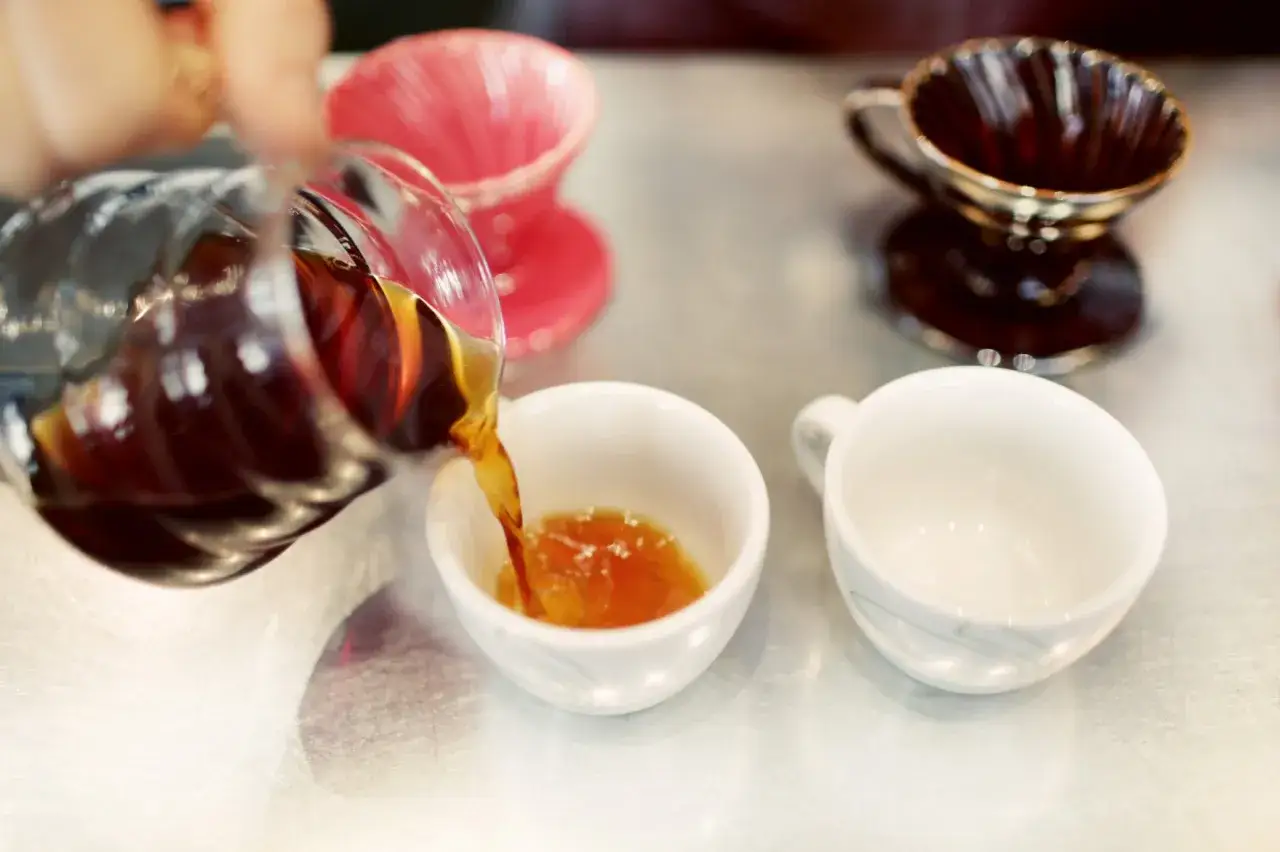 Här hälls nybryggt arabica kaffe i en vit kopp. Två andra koppar och två filterhållare syns i bakgrunden.