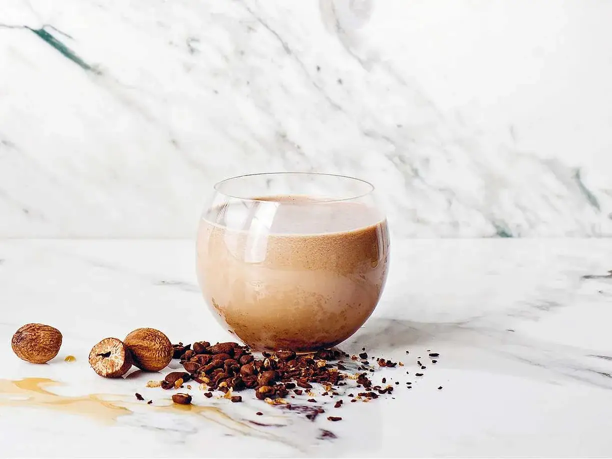En krämig chai kaffe-drink i ett glas, med nötter och kaffebönor utspridda på en marmoryta.