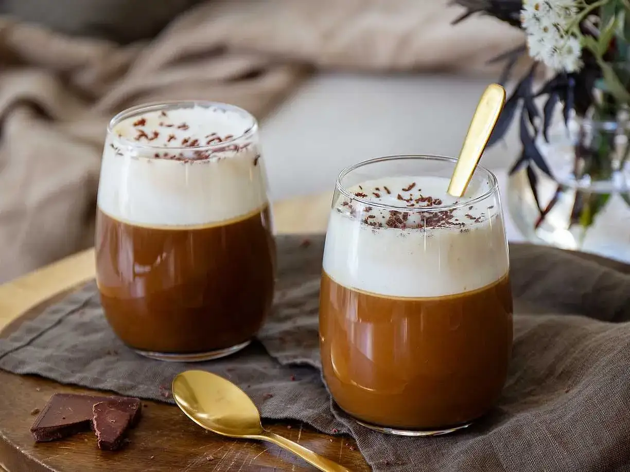 Två glas med kaffe Karlsson, toppade med vispgrädde och chokladströssel. En gyllene sked vilar i det ena glaset.