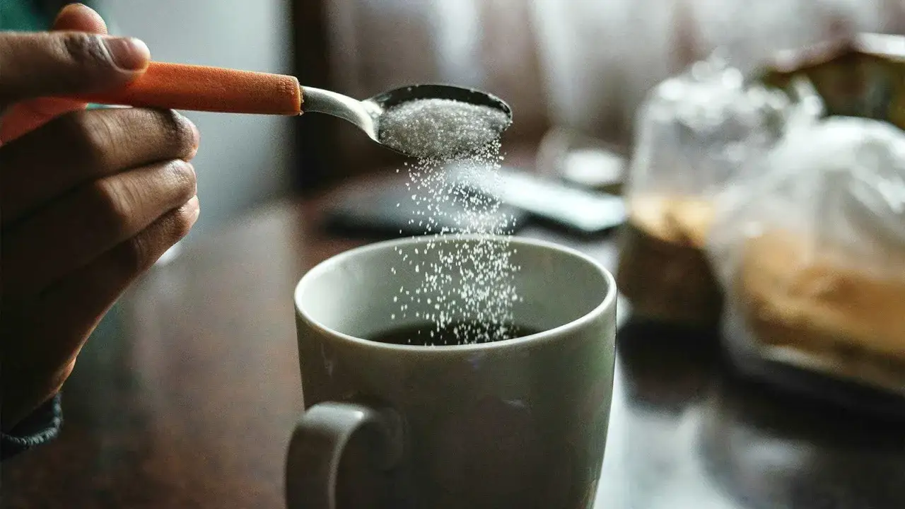 En hand häller socker från en sked ner i en kaffekopp. Kanske en liten mängd salt i kaffet för att framhäva smaken?