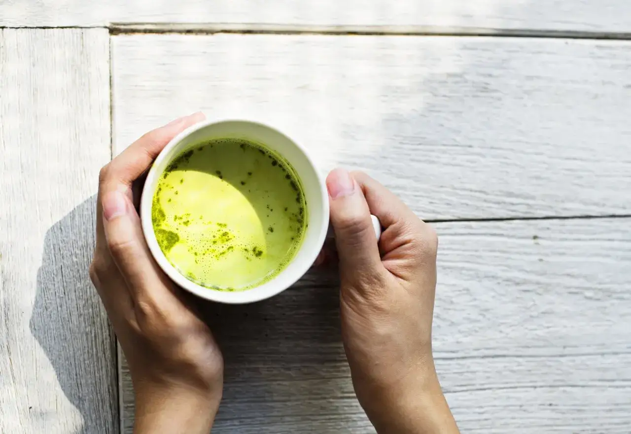 Händer håller en kopp med grön matcha, en dryck rik på matcha koffein, på ett vitt träbord.