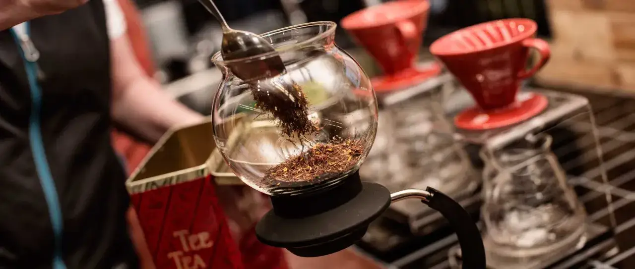 En sked lägger teblad i en glaskaraff, redo att brygga det godaste teet. Röda kaffefilter står i bakgrunden.