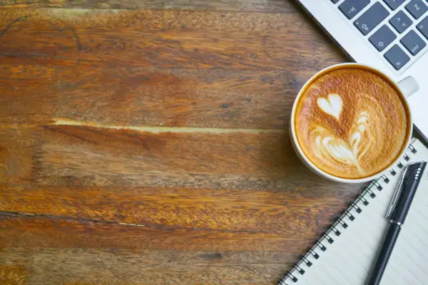 En kopp kaffe med lattekonst, en laptop och en anteckningsbok på ett träbord. Perfekt för att få lite koffein och inspiration.