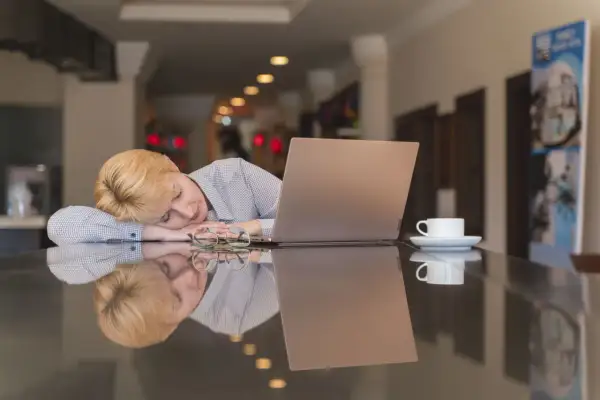 Kvinnan med blont hår sover vid ett bord med en laptop och en kaffekopp. Hon verkar trött, trots att hon inte blir pigg av kaffe.