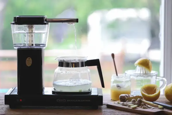 Vatten droppar från en kaffebryggare ner i en glaskanna, redo att brygga te. Ingredienser som citron och ingefära syns i bakgrunden.