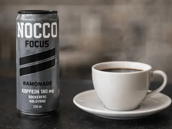 En burk NOCCO Focus Ramonade med 180 mg koffein bredvid en ångande kopp kaffe. Perfekt för att kickstarta dagen med extra espresso koffein.