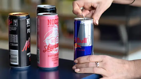 En hand öppnar en burk Red Bull, bredvid NOCCO-burkar. Koffein redbull för energi.