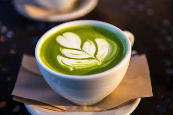 En vit kopp med grön matcha latte, dekorerad med ett vitt mönster som liknar ett löv. Matcha innehåll i sin finaste form.