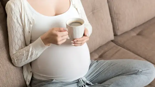 En gravid kvinna sitter på en soffa och håller en mugg med kaffe.