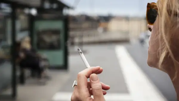 En kvinna röker en cigarett vid en busshållplats. Bilden kan illustrera prisutveckling cigaretter.